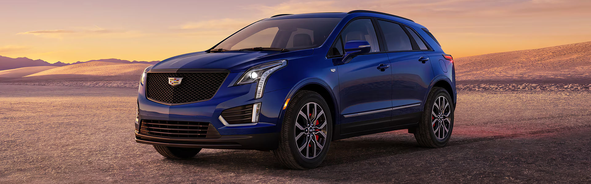 Blue Cadillac XT5 parked on a desert road at sunset, featuring a bold front grille, sleek LED headlights, and a scenic landscape in the background.